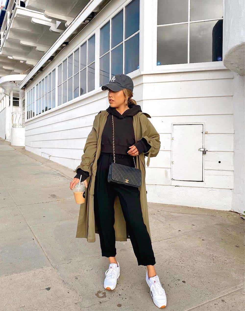 Woman wearing black pants, a black cropped hoodie, white sneakers and a green trench coat.