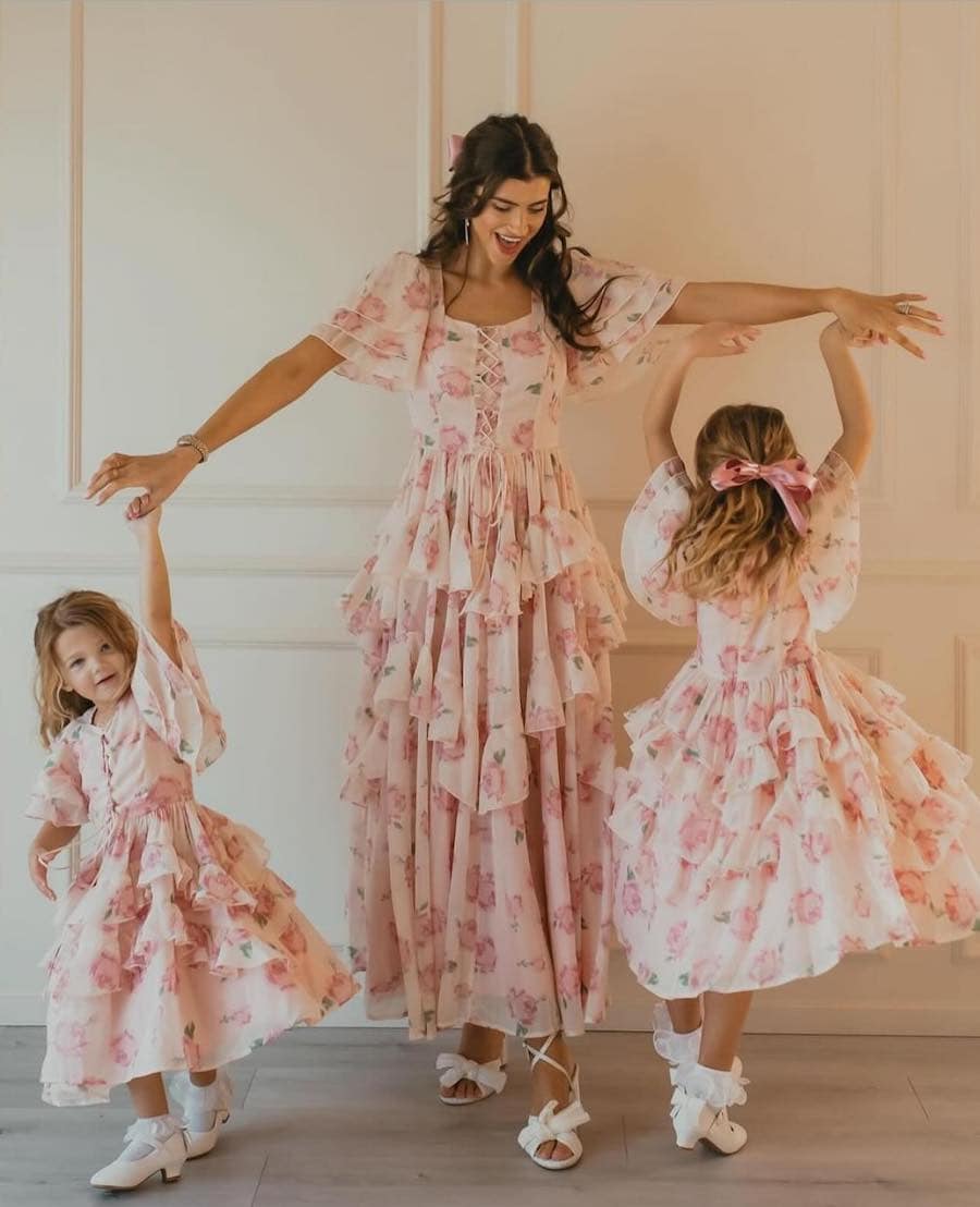 A spring family photo with a mom and her two daughters wearing matching dresses featuring a tiered ruffled skirt, pink floral print, and a lace up front