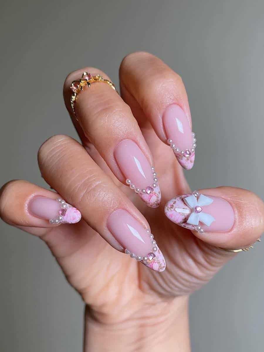 Long nude almond nails with pink romantic art French tips bordered by pearls, pink heart-shaped gems, and accented with a white bow charm