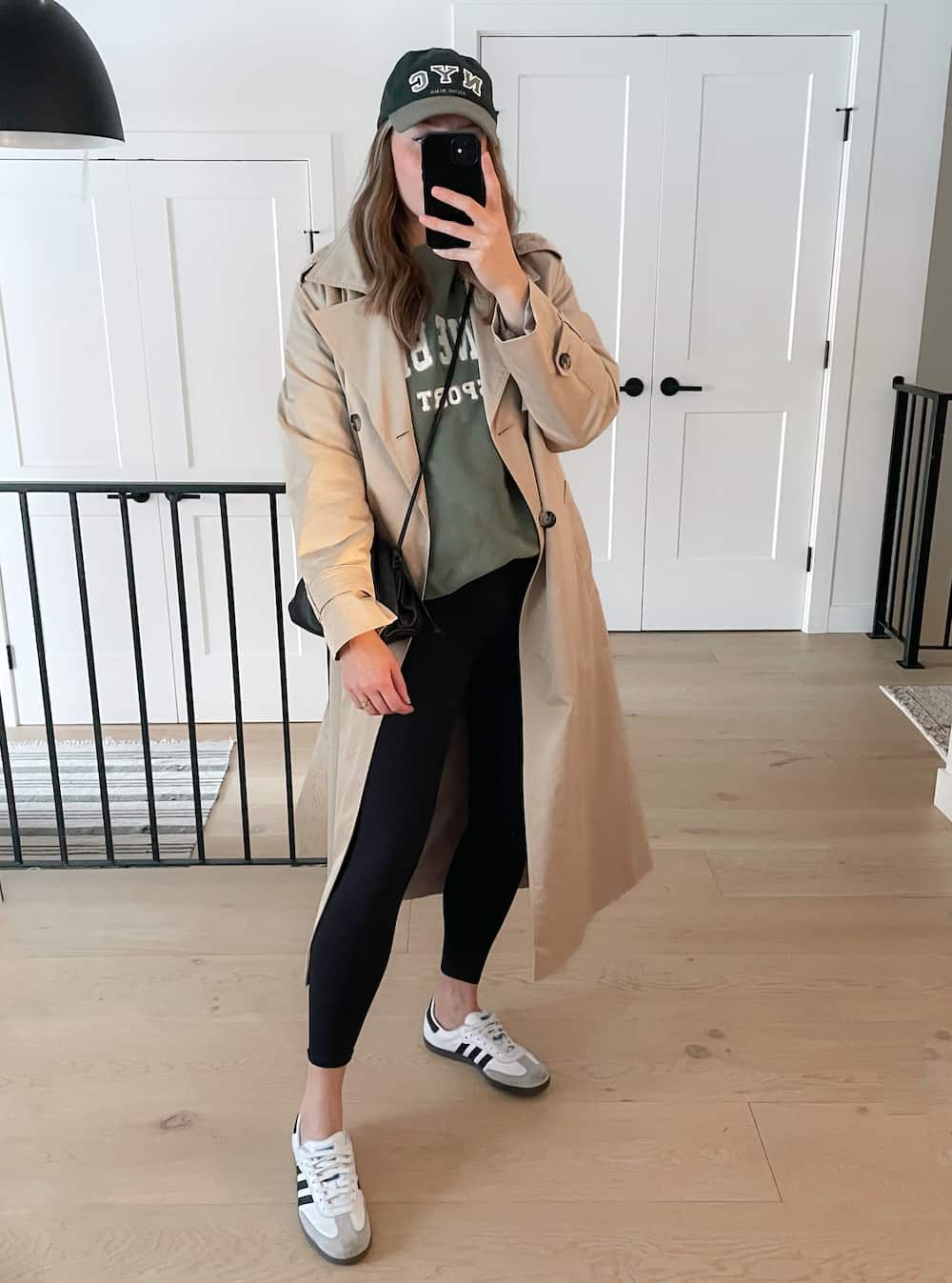 Christal wearing black leggings, an olive sweatshirt and a tan trench coat with sneakers and a baseball cap.