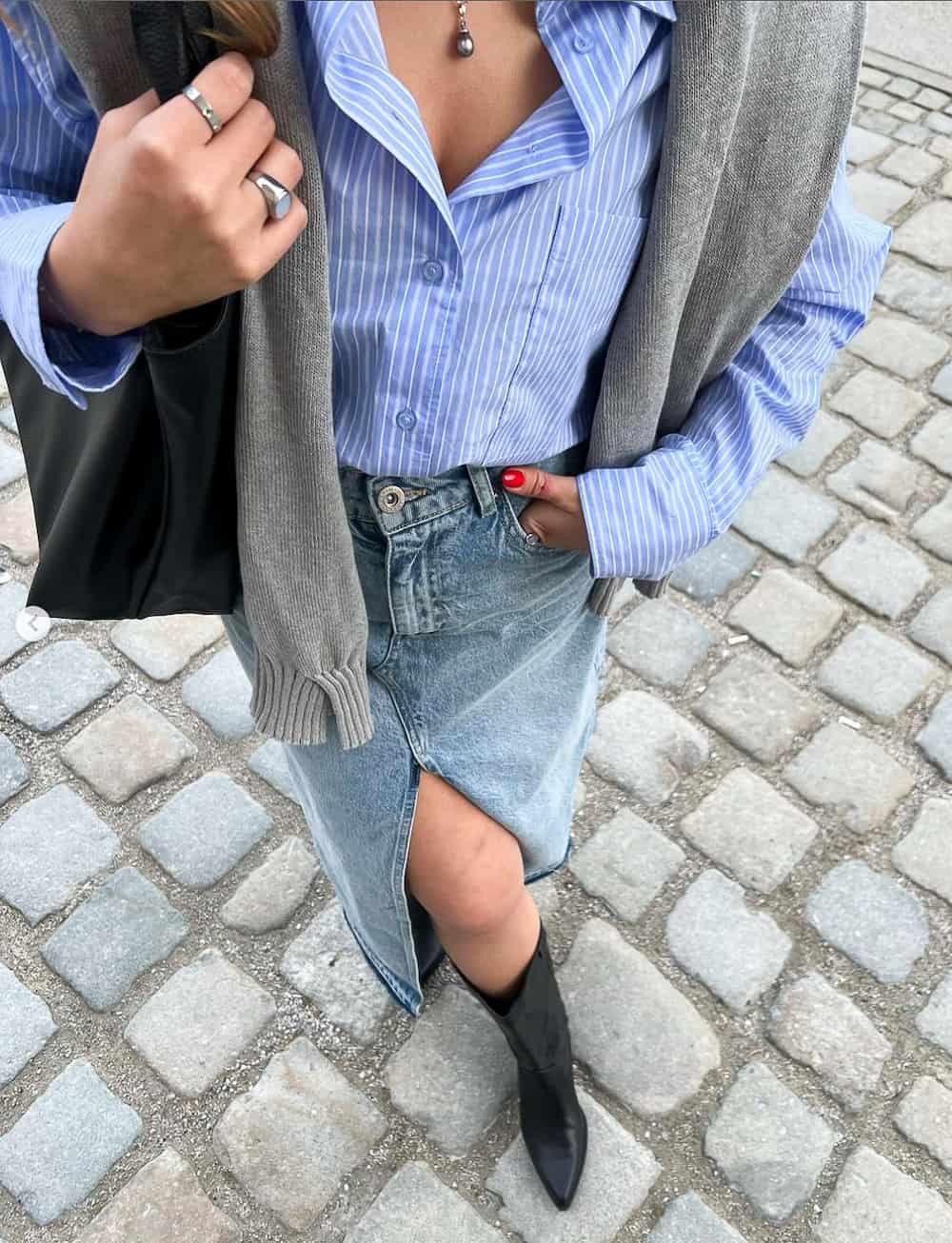 Woman wearing a denim maxi skirt with a blue and white striped button down, a grey cardigan and black booties.