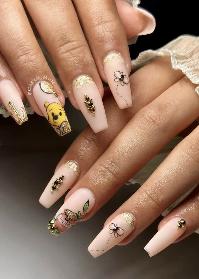 Long coffin nails with a Winnie the Pooh design featuring matte nude nails, gold details, and nail art