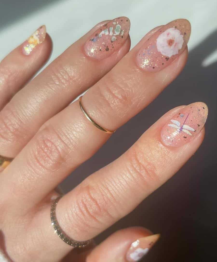 Short almond nails with nude polish topped with chunky glitter polish, floral accents, and dragonfly nail stickers