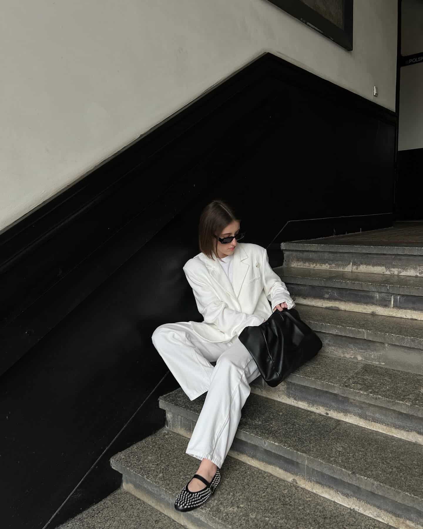 woman sitting on steps wearing a white oversized blazer with white trousers and studded black mary jane flats