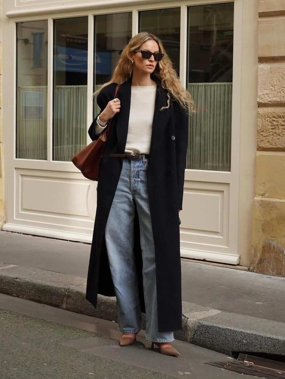Woman wearing baggy jeans, pointed toe pumps, a cream sweater and an oversized black trench coat.