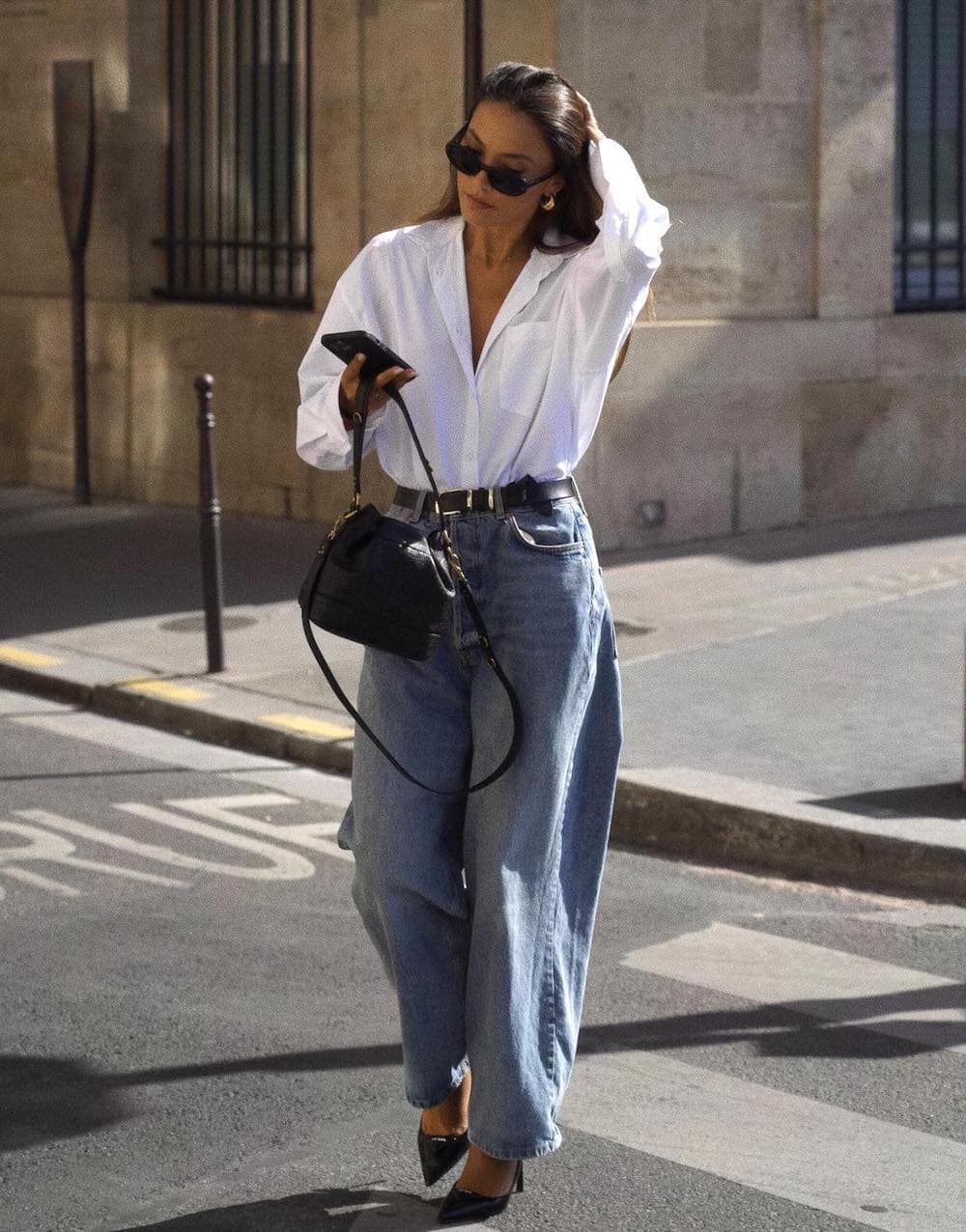 Woman wearing baggy jeans, black pointed toe pumps, a white oversized button down and black accessories.