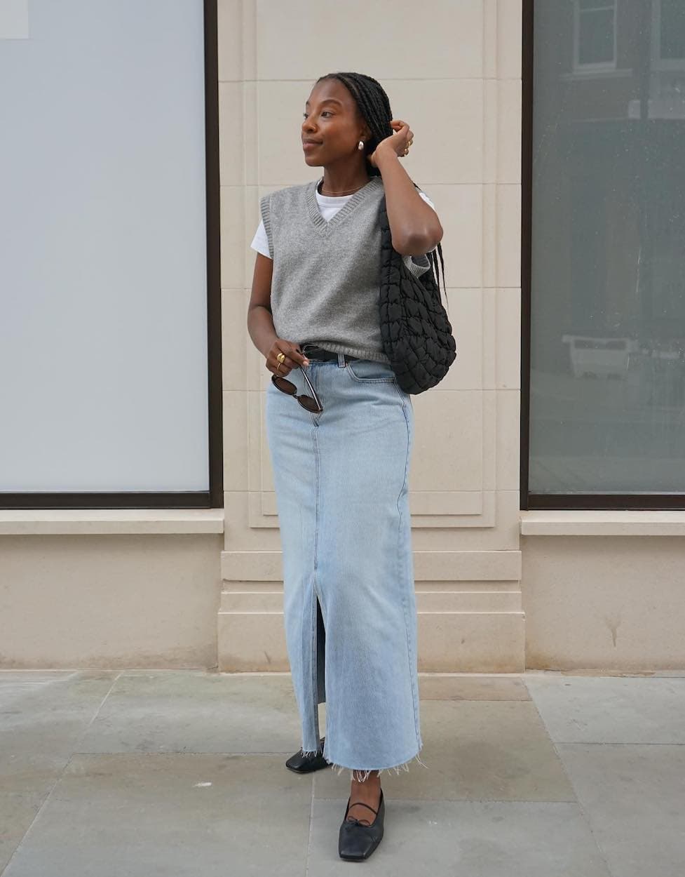 Woman wearing a denim midi skirt with a whit t-shirt, a grey sweater vest and black ballet flats.