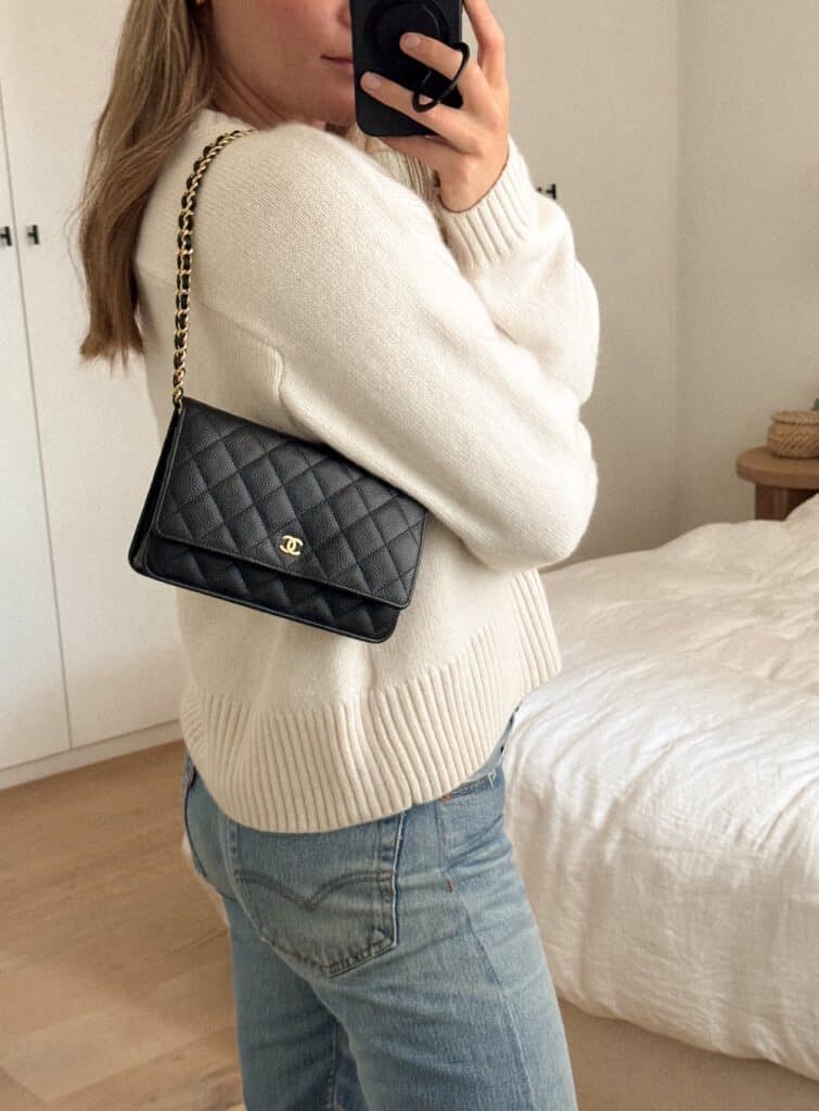 Woman wearing a cream sweater with a black Chanel wallet on chain over her shoulder and blue jeans