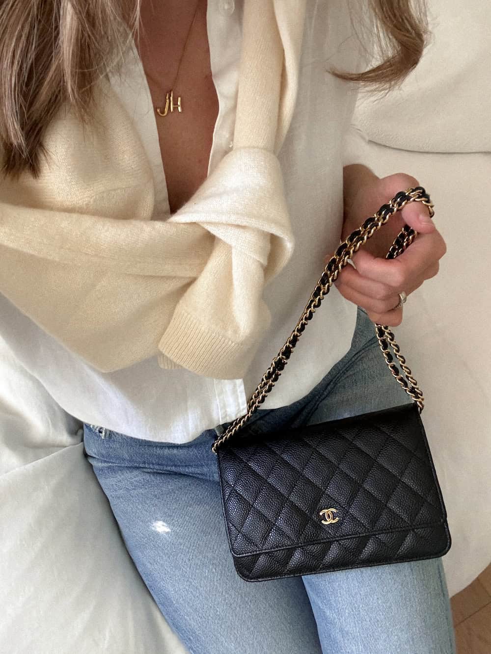 Woman holding a black Chanel wallet on chain wearing a white button up shirt, cream sweater, and blue jeans