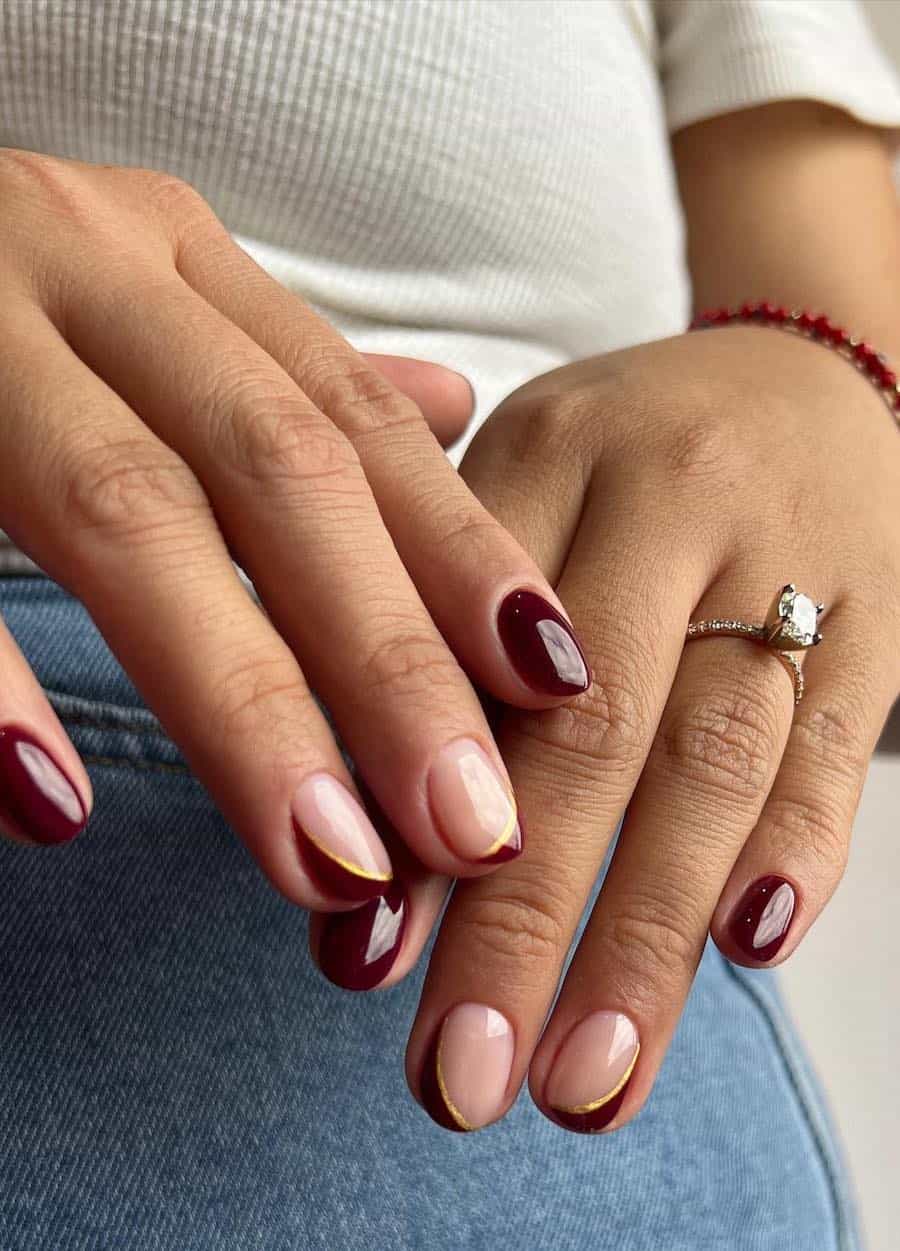 short squoval nails painted wine red with two glossy nude accent nails featuring asymmetrical tips with gold borders