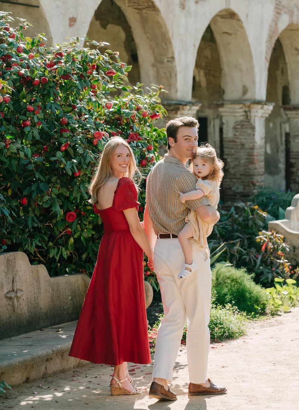 a summer family photo shoot where the mom is wearing a crimson dress, while the father and daughter wear classic neutral pieces