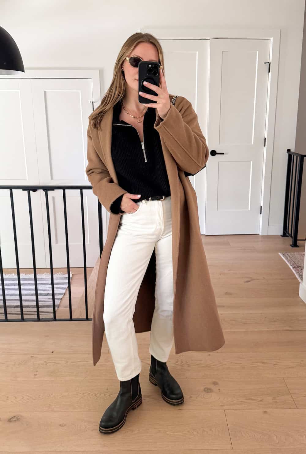 Christal wearing white jeans, a black quarter zip sweater, black boots and a long camel colored coat.