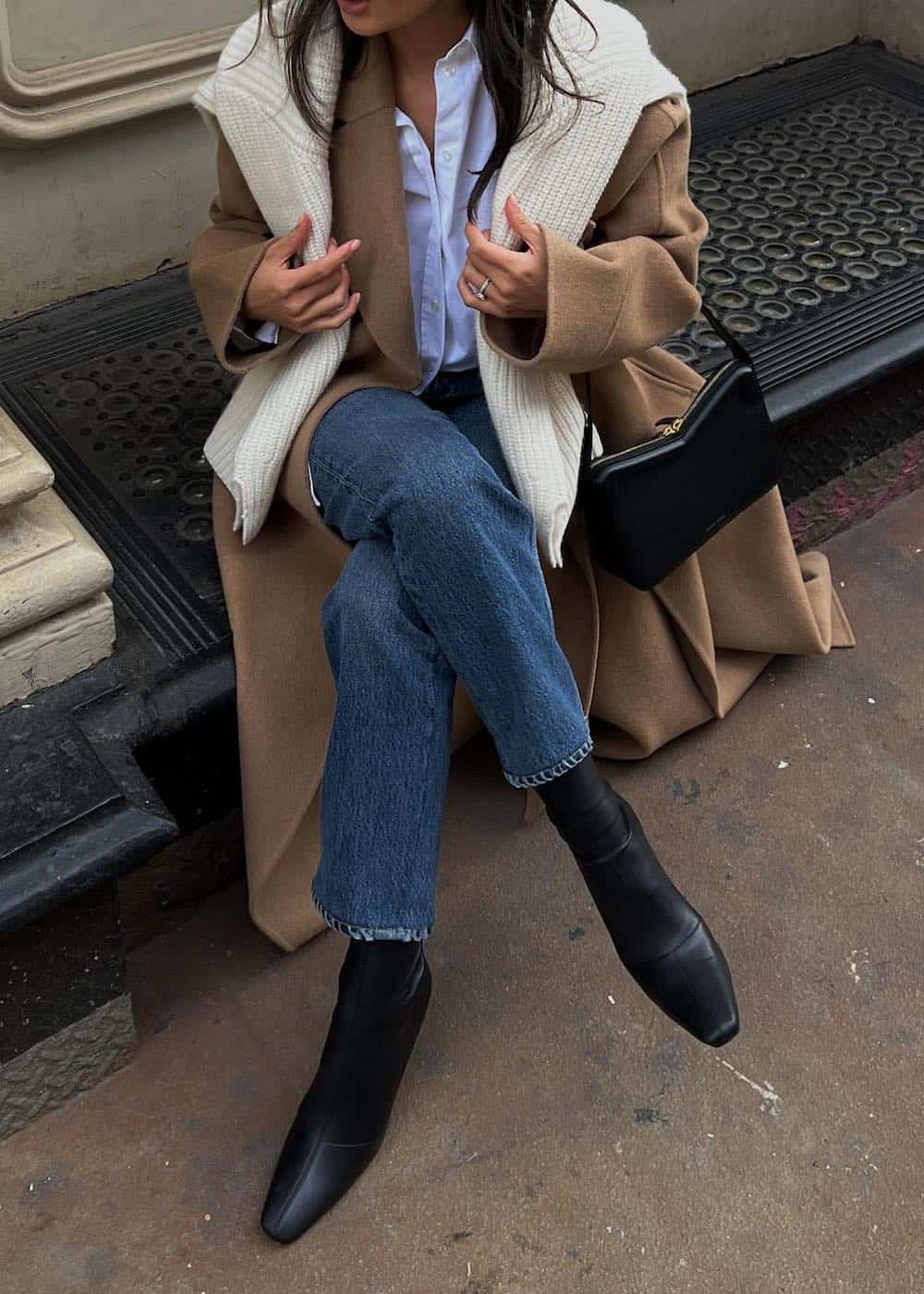 Woman wearing dark denim pants, black booties, a white button down, a camel colored trench coat and a white sweater over her shoulders.