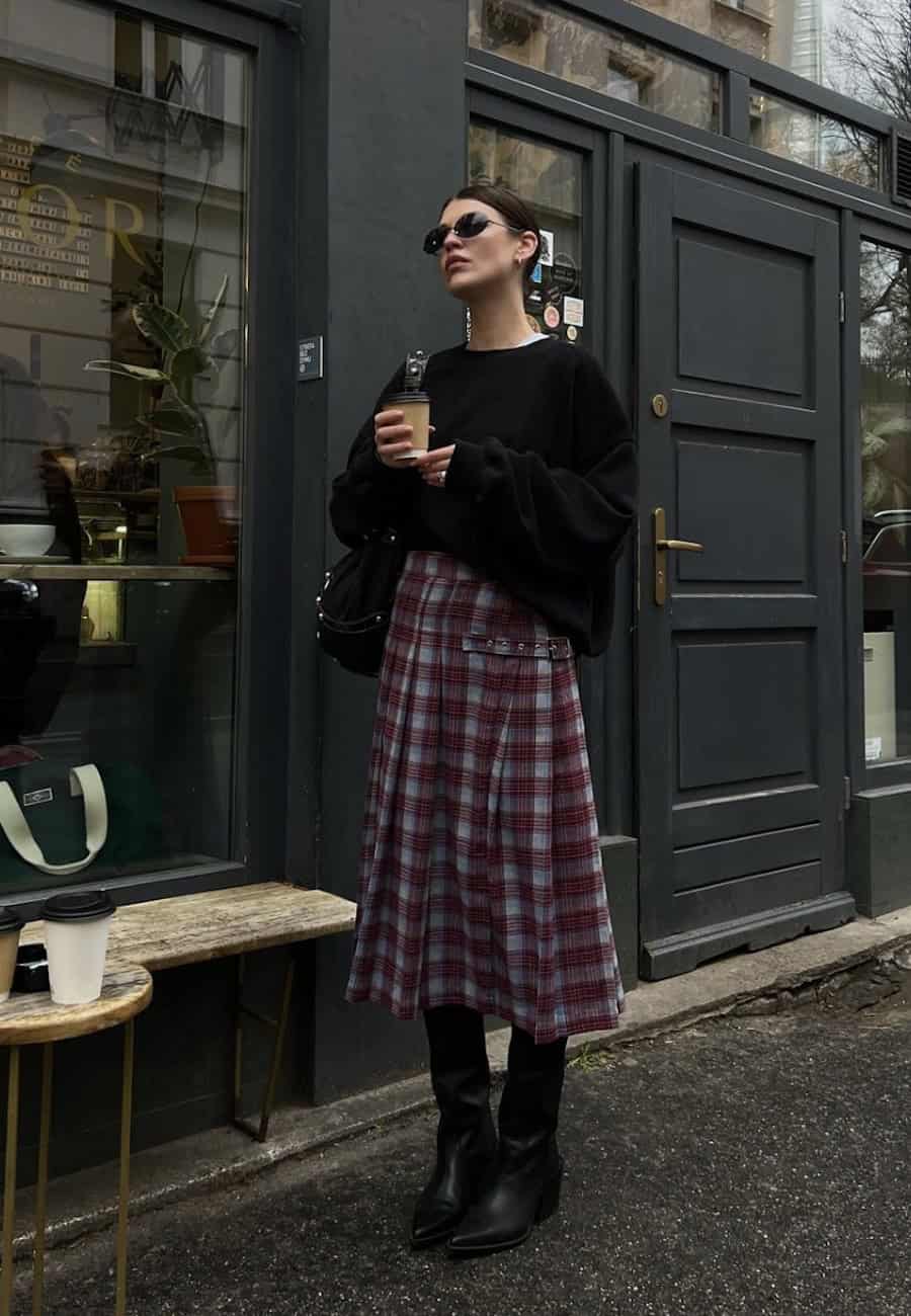 Woman wearing a plaid midi skirt with a black oversized sweater and black boots.