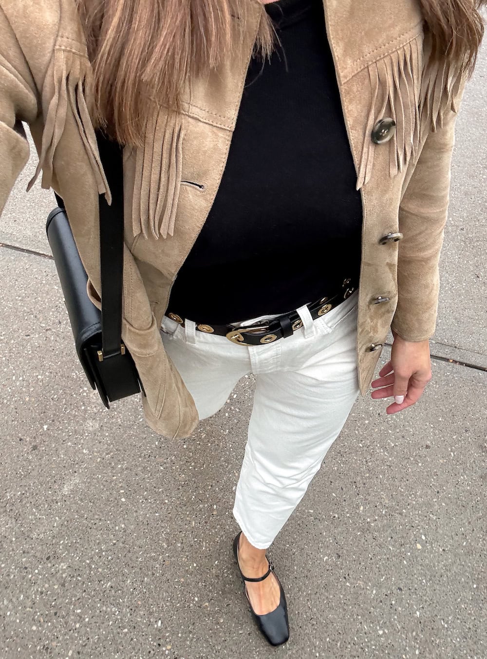 woman wearing a tan fringe suede jacket over a black t-shirt with off-white denim, a black leather belt with gold grommets, and black Mary Jane flats