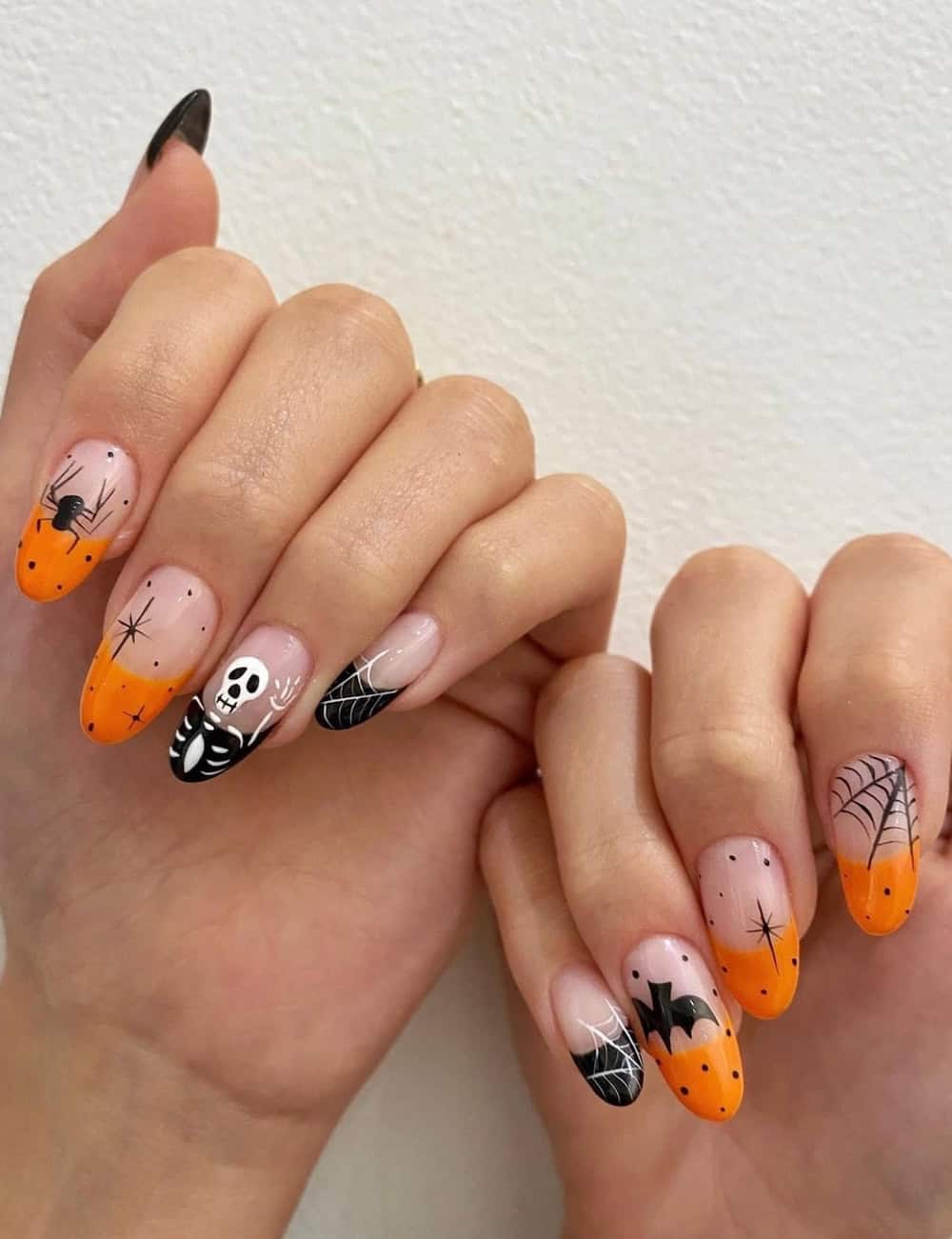 long nude almond nails with black and orange tips and Halloween nail art featuring skeletons, spiders, and bats