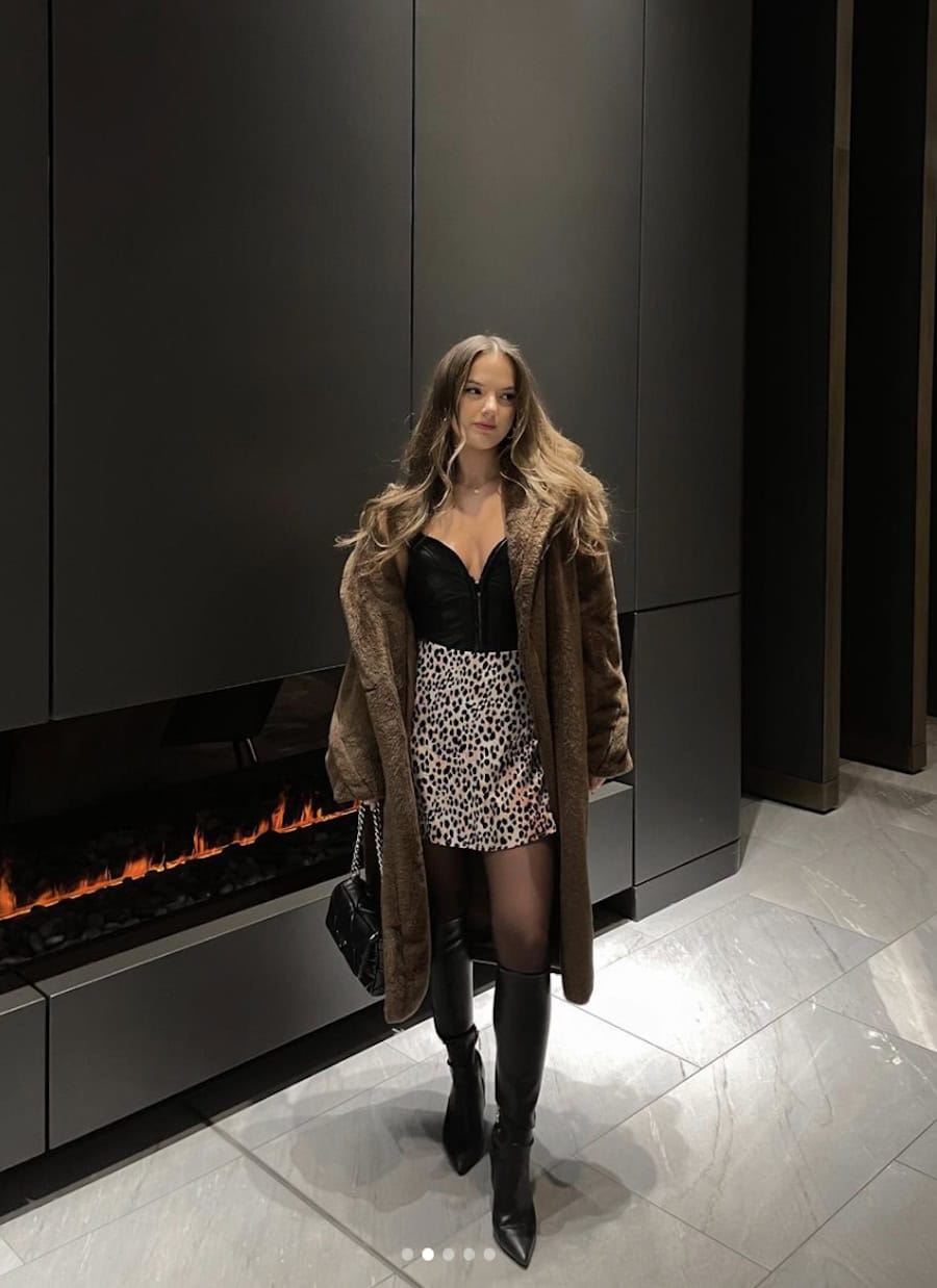 a winter concert outfit featuring tall black boots, an animal print mini skirt, a black corset top, and a long brown faux fur coat