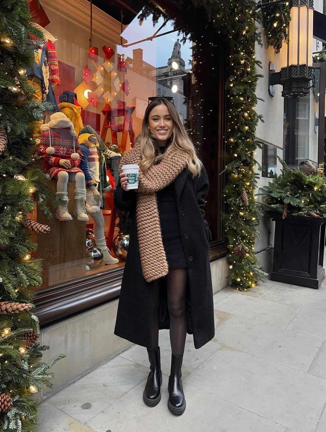 Woman wearing a black mini dress with black sheer tights, black lugg boots, an oversized black coat and a brown scarf.