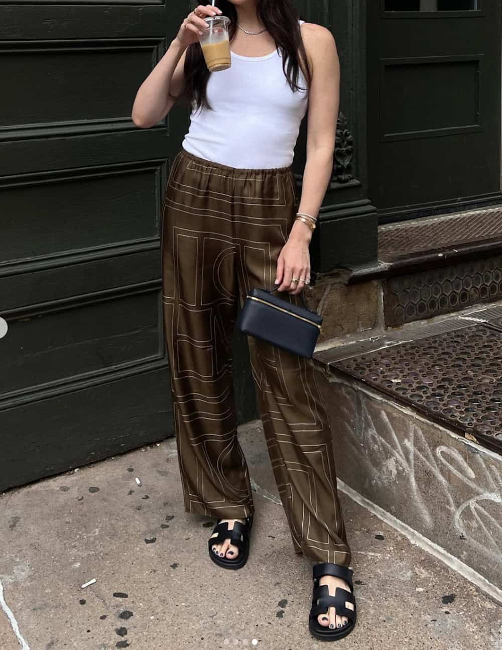 Woman wearing brown silk pants with a geometric pattern and a white tank top with black chunky sandals.