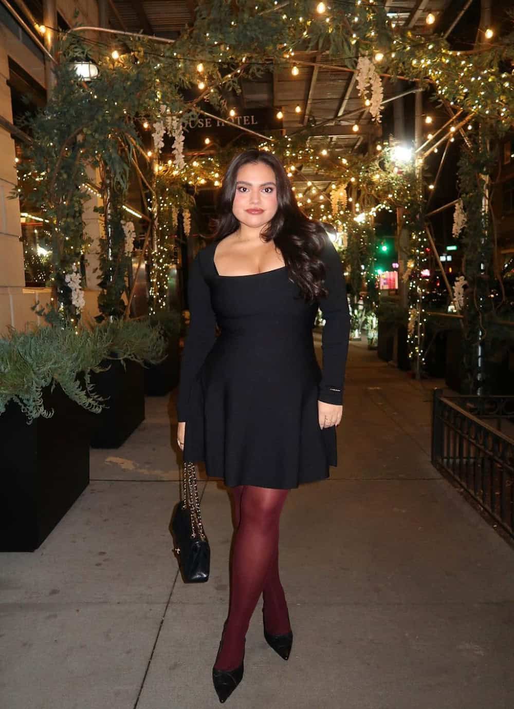 Woman wearing a long sleeve black dress with red tights and black pumps.