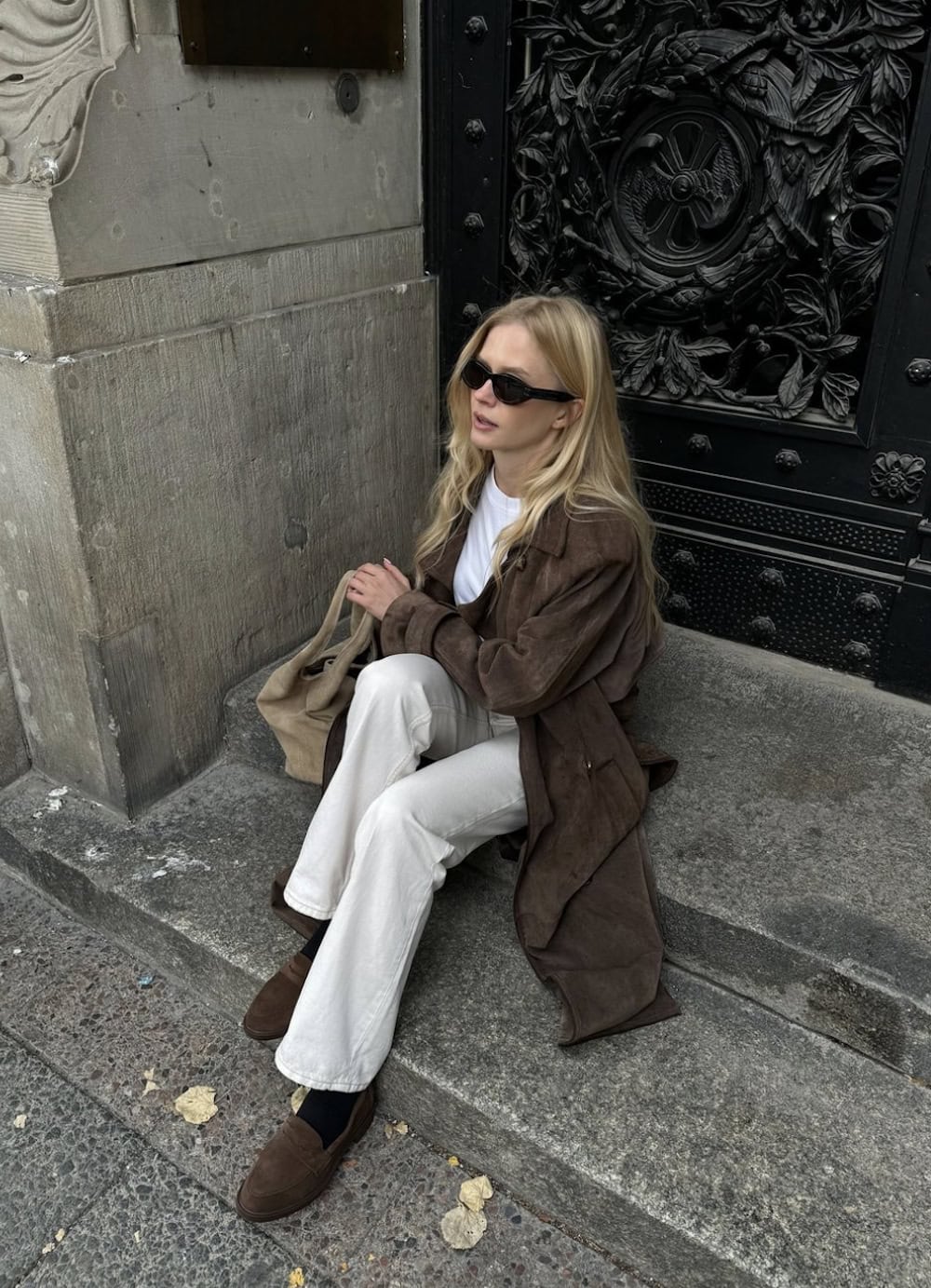 Woman wearing white jeans with a long brown coat and brown loafers.