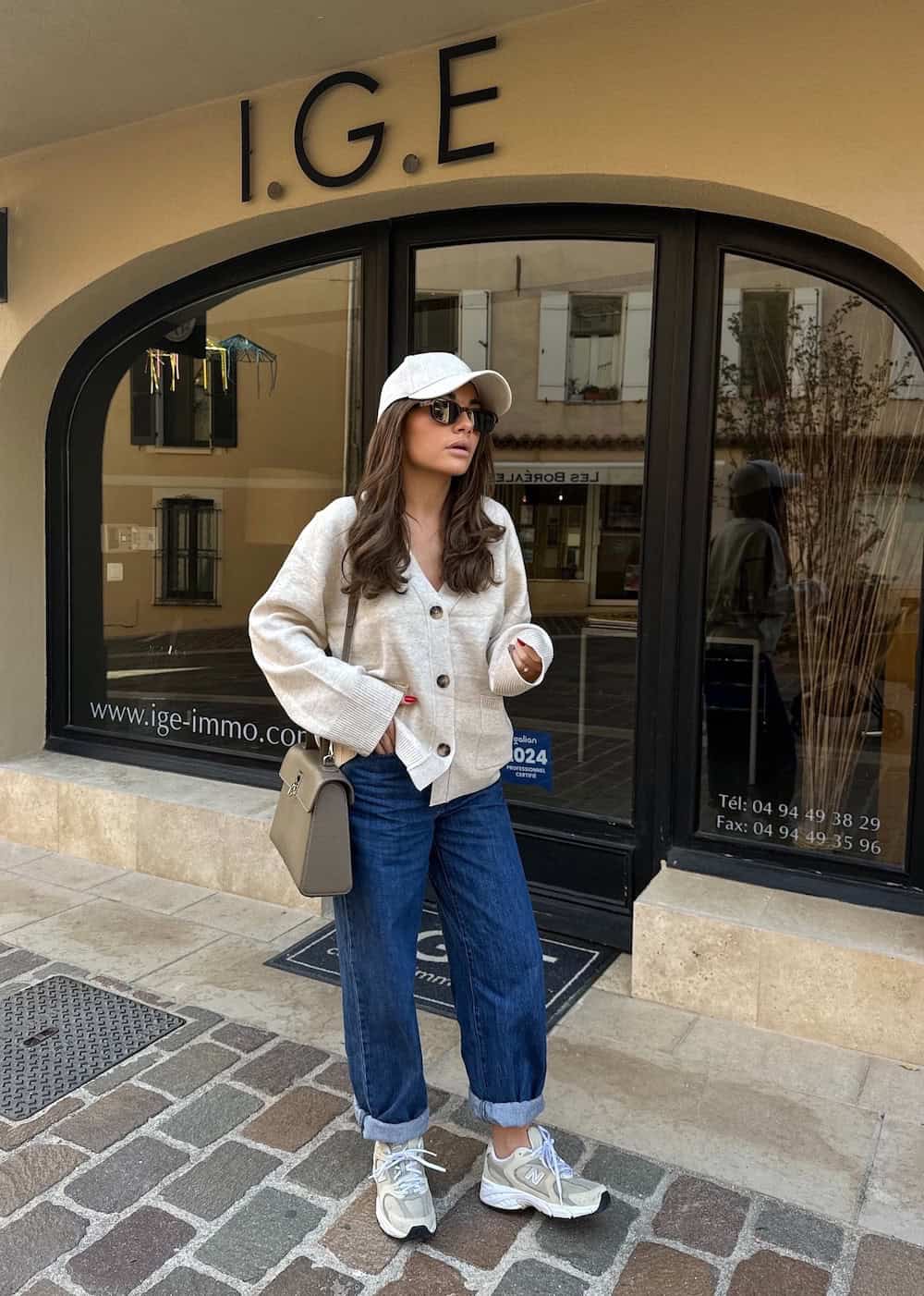 Woman wearing rolled up jeans with New Balance sneakers and a white cardigan with a baseball cap.