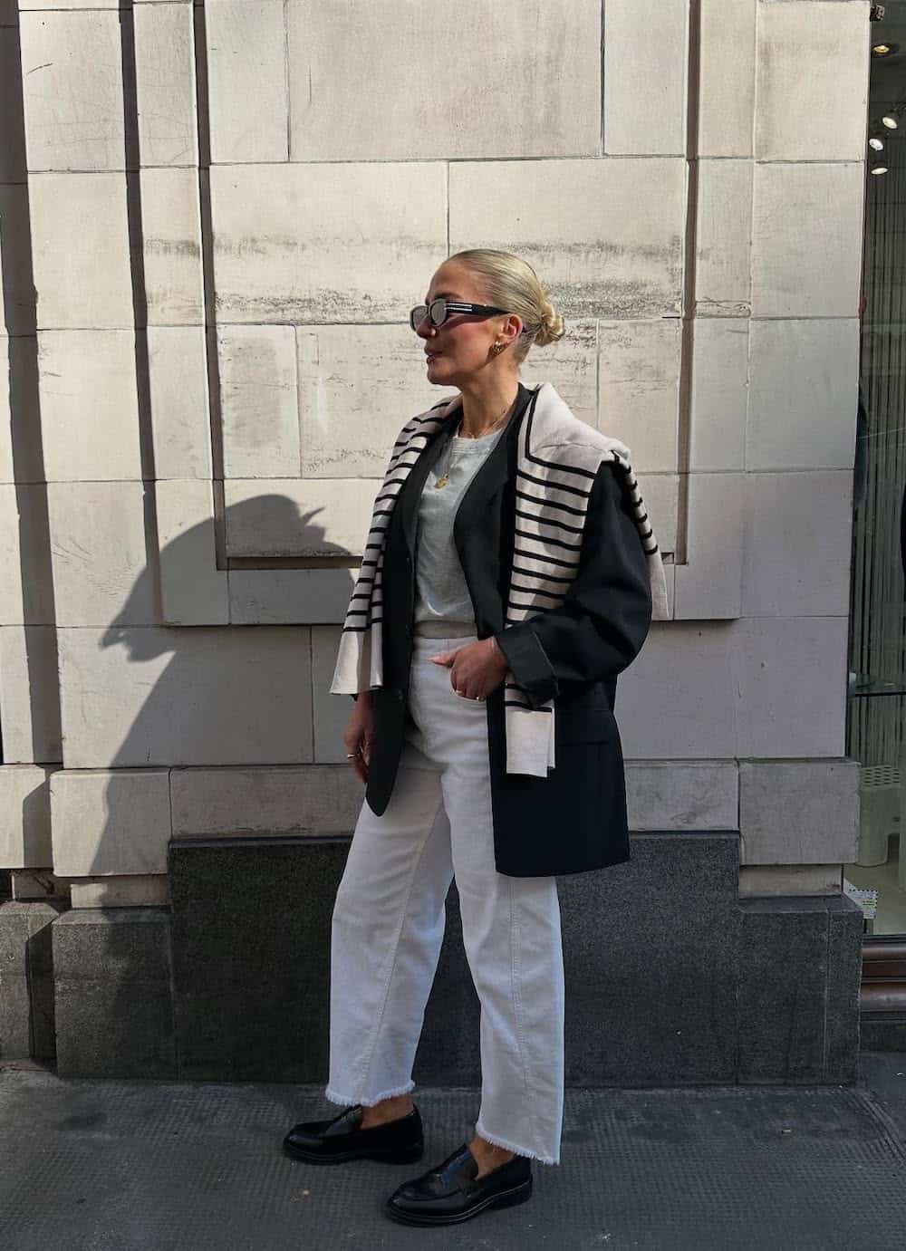 a woman wearing white jeans, a grey tee, a black blazer, a striped sweater over the shoulders, and black leather loafers