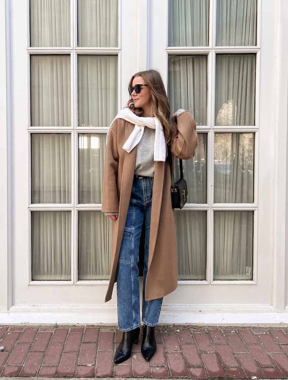 a woman wearing blue jeans, a grey sweater, a long brown coat, and pointed black boots