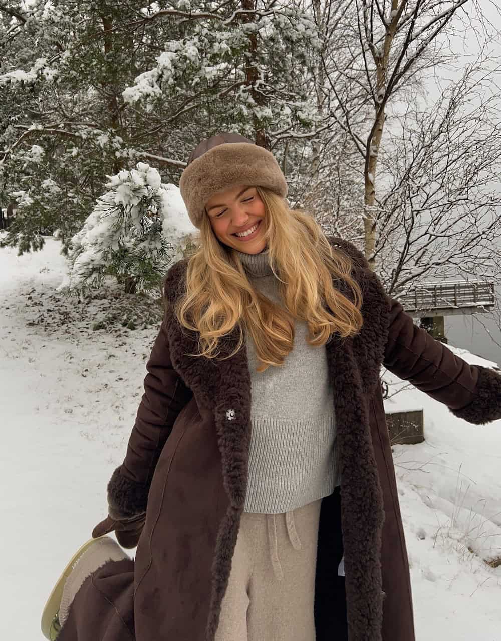 a woman wearing beige sweatpants, a grey turtleneck, an oversized brown coat, and a brown beanie
