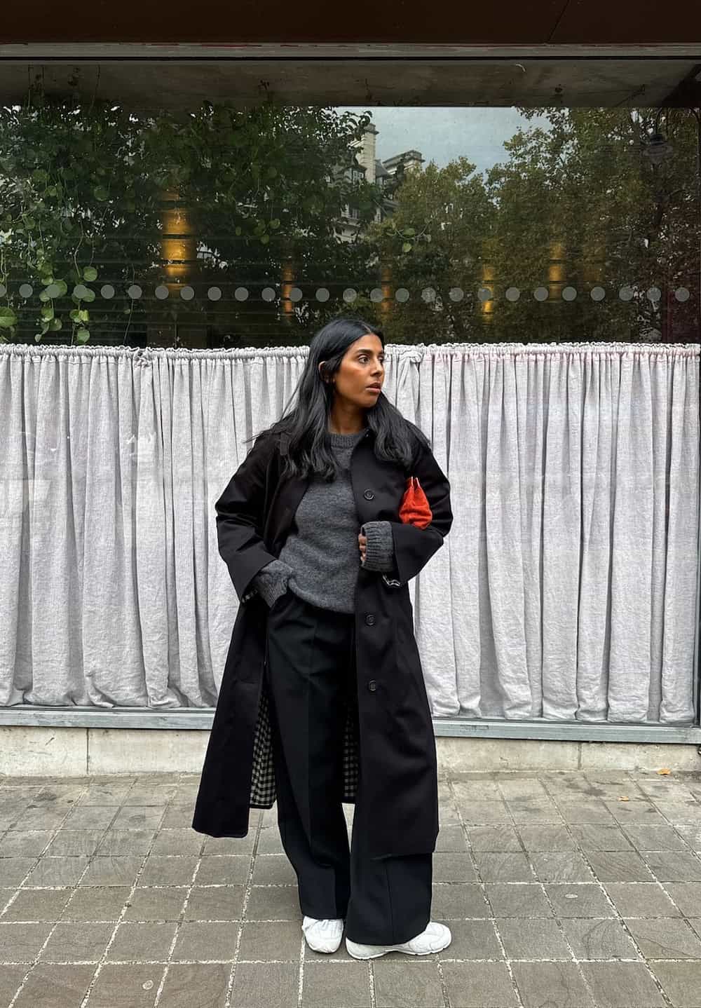 a woman wearing black pants, a grey sweater, a long black coat, and white sneakers