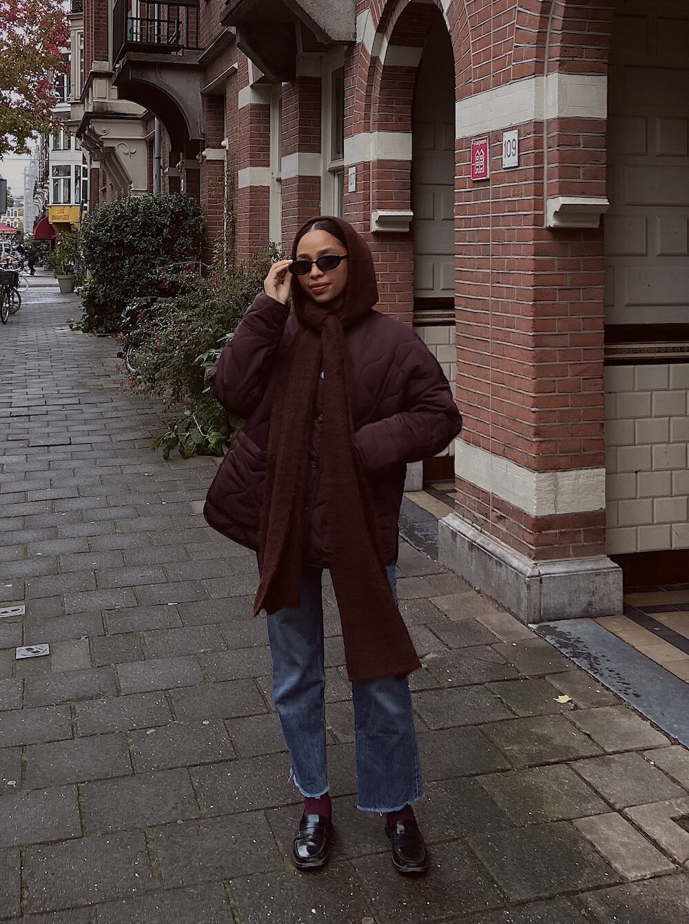a woman wearing blue jeans, a brown quilted coat, a long brown scarf, and black loafers