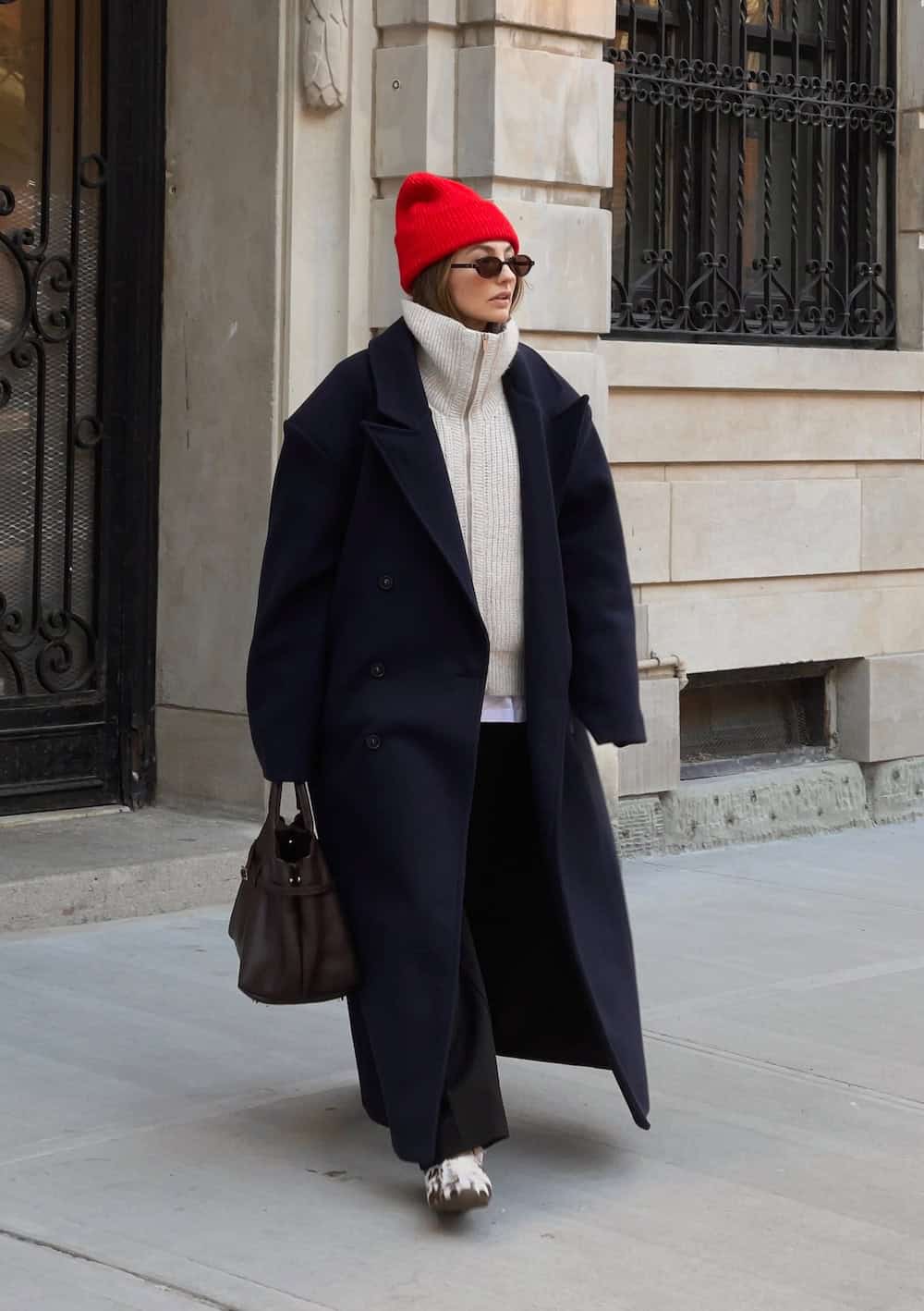 a woman wearing black trousers, an ecru half zip, a long black coat, and a red beanie