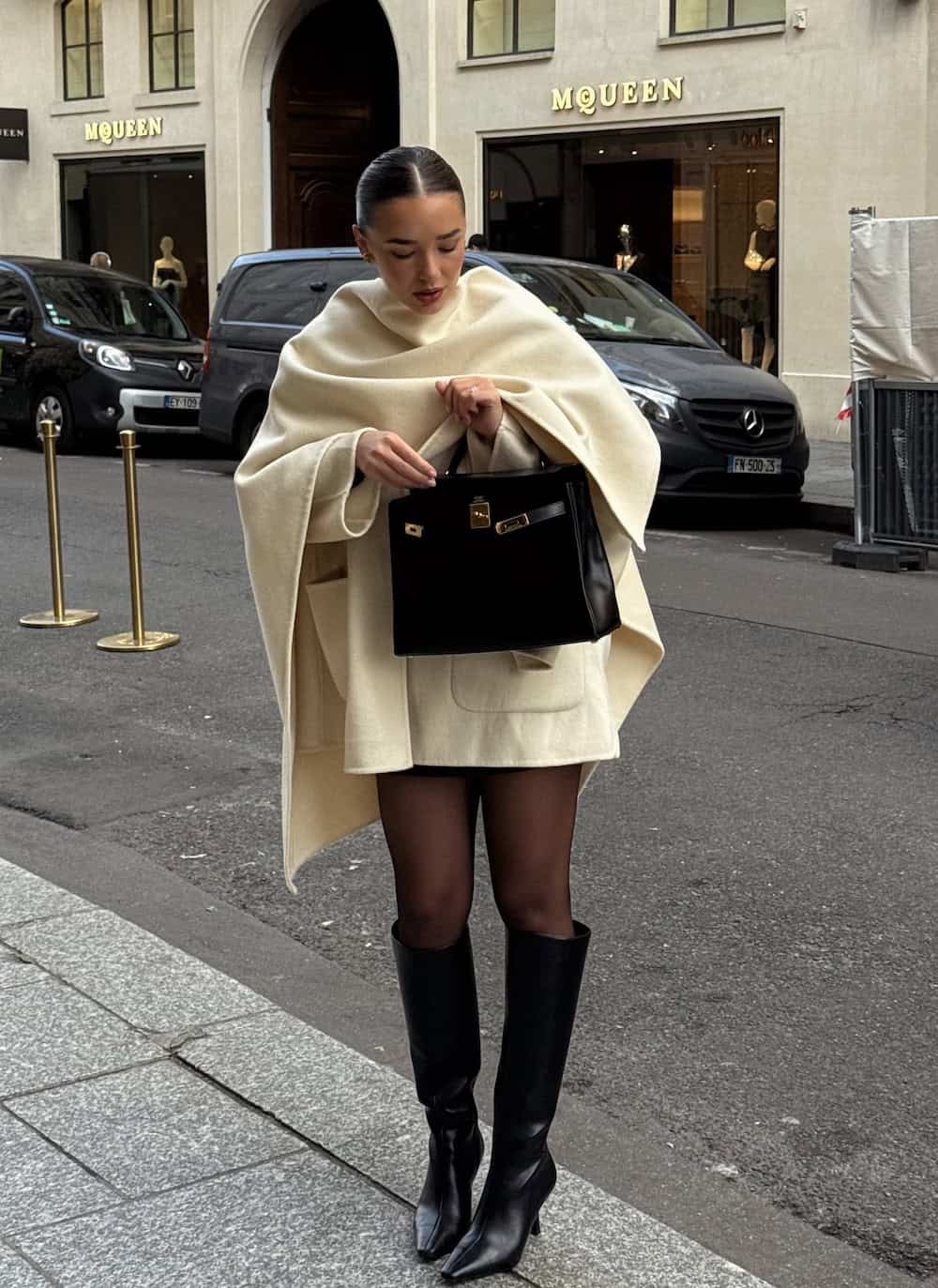 a woman wearing sheer tights, knee high boots, and a mini cream-colored dress with a cape