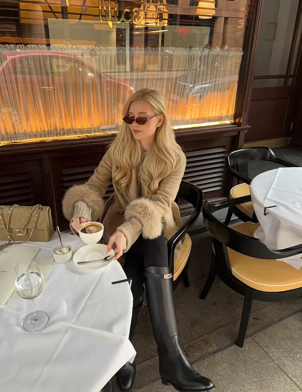 a woman wearing black leggings, a fur trimmed beige coat, and tall black boots