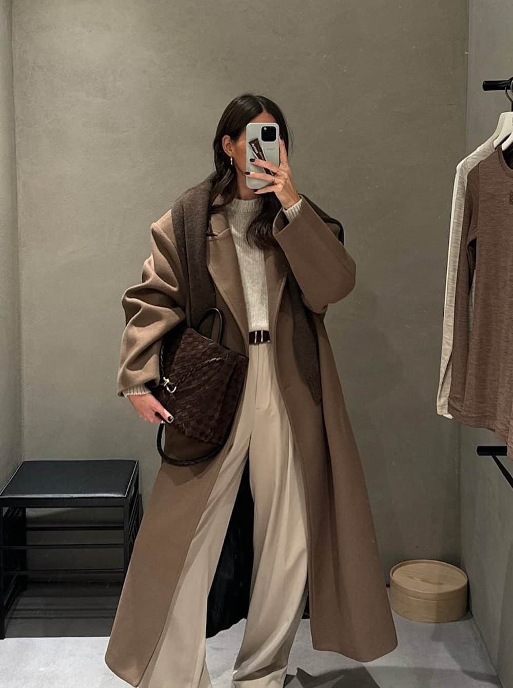 a woman wearing light beige trousers, a knit crewneck sweater, and a long light brown overcoat