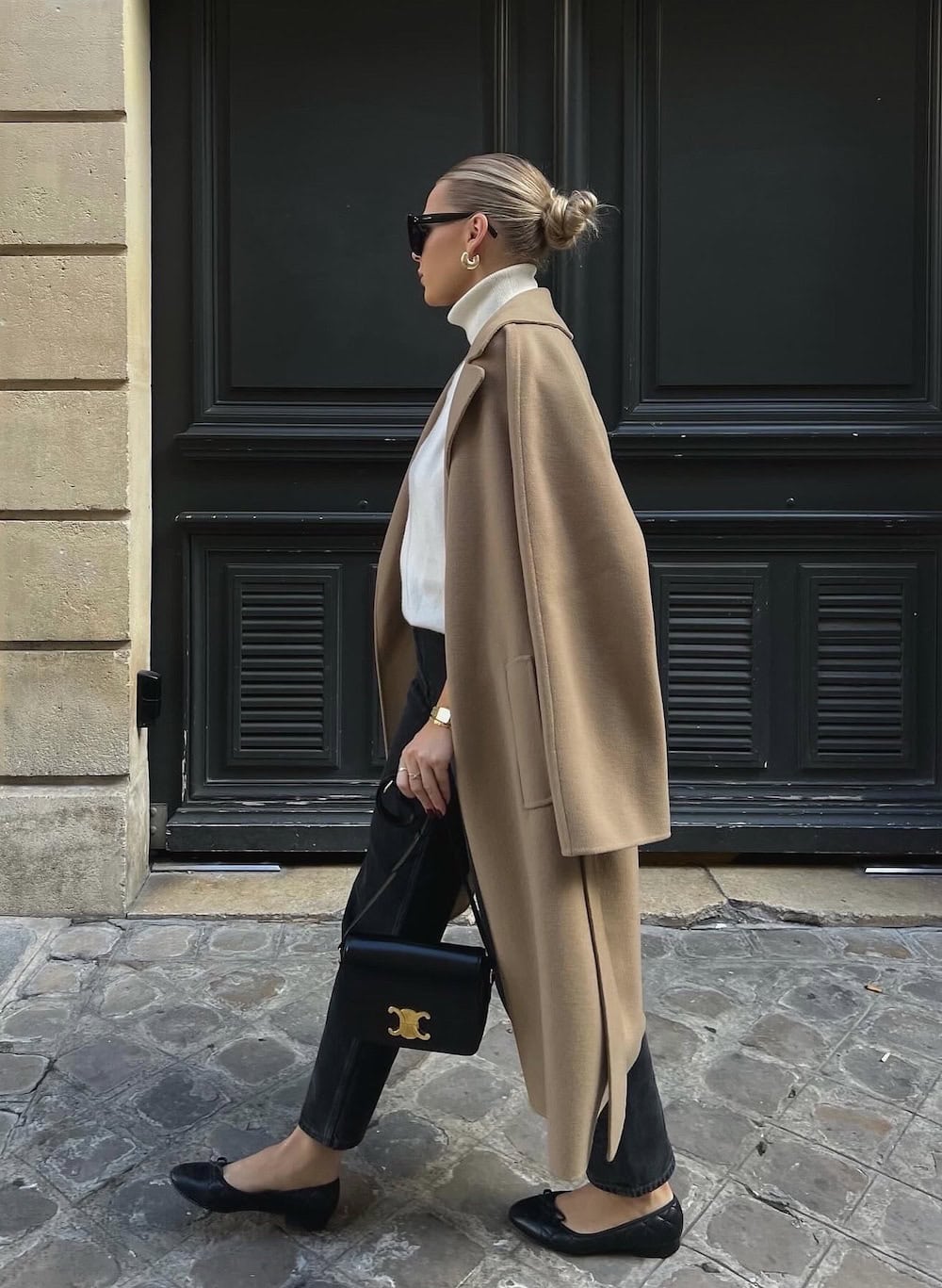 a woman wearing black jeans, a white turtleneck, a long beige coat, and black ballet flats