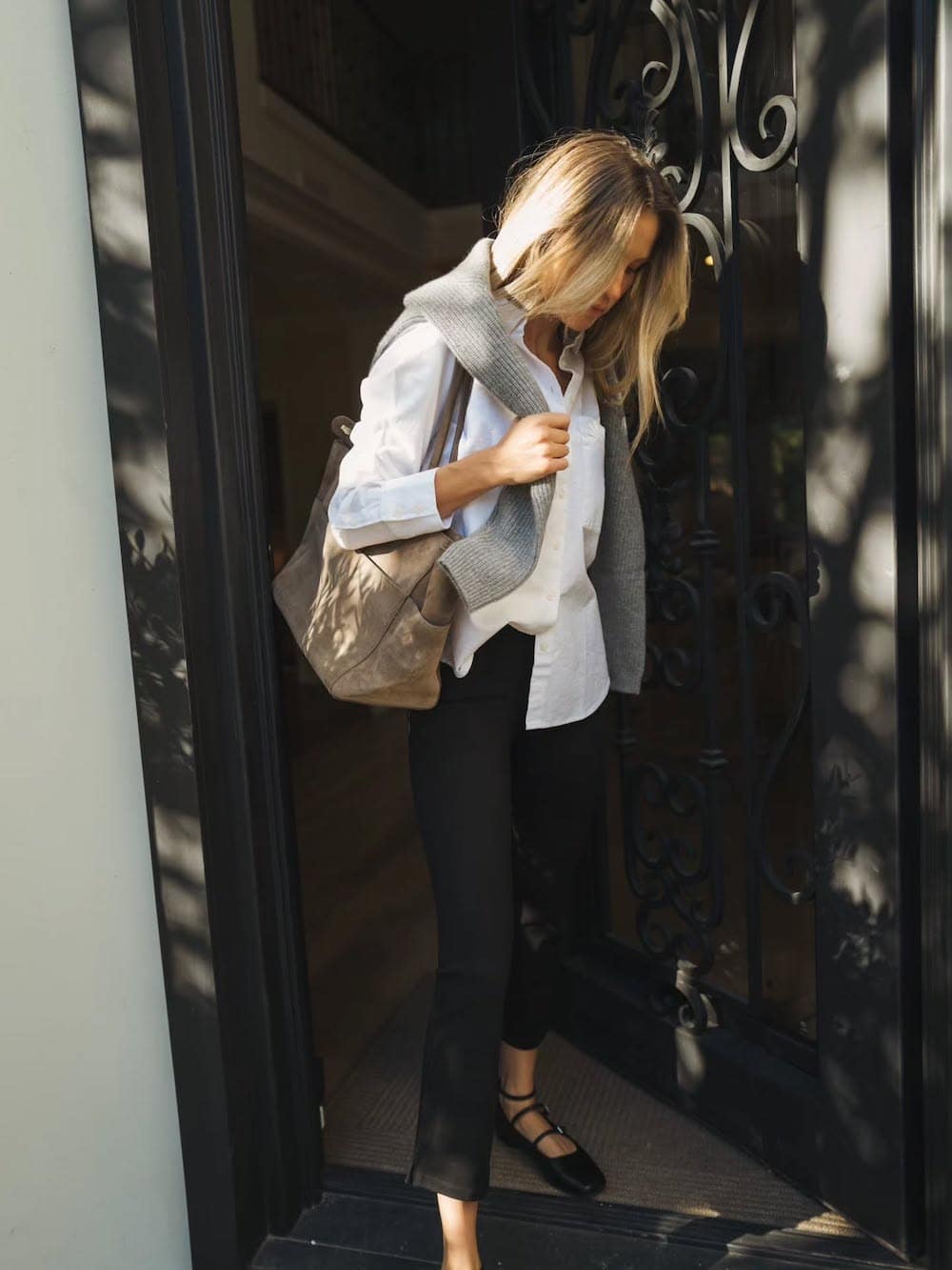a woman wearing black capri pants, a white button up, a grey over the shoulder sweater, and mary jane ballet flats